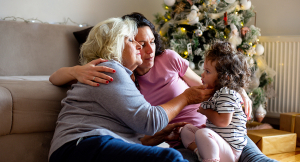 A young girl enjoys the attention her mother and grandmother are giving to her. They are on the floor, in the living room.